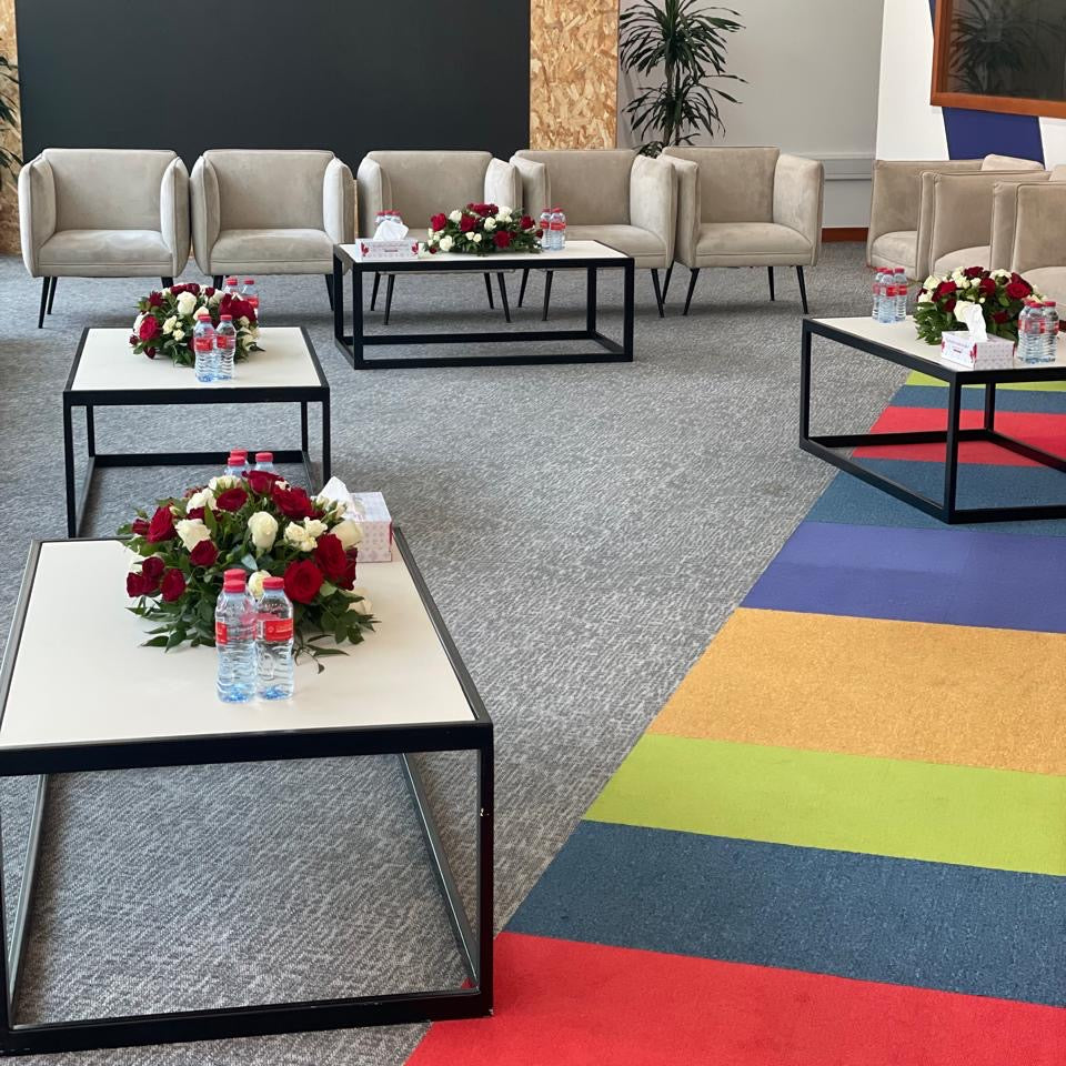 Modern office lobby with beige sofas, coffee tables, and flower arrangements on a multicolored carpet.