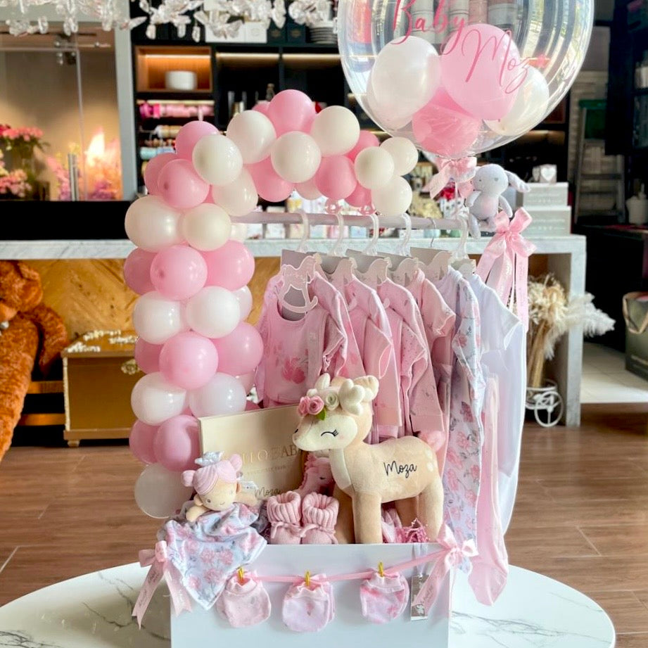 Decorative setup with pink and white balloons, baby clothes, and toys on a stand.