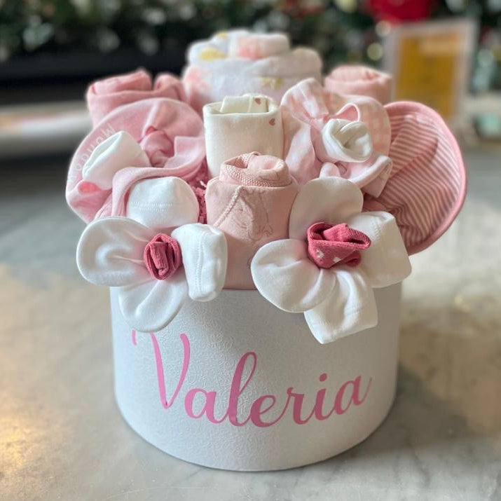Decorative flower arrangement in a white container with 'Valeria' on it, placed on a marble surface with a blurred Christmas tree in the background.