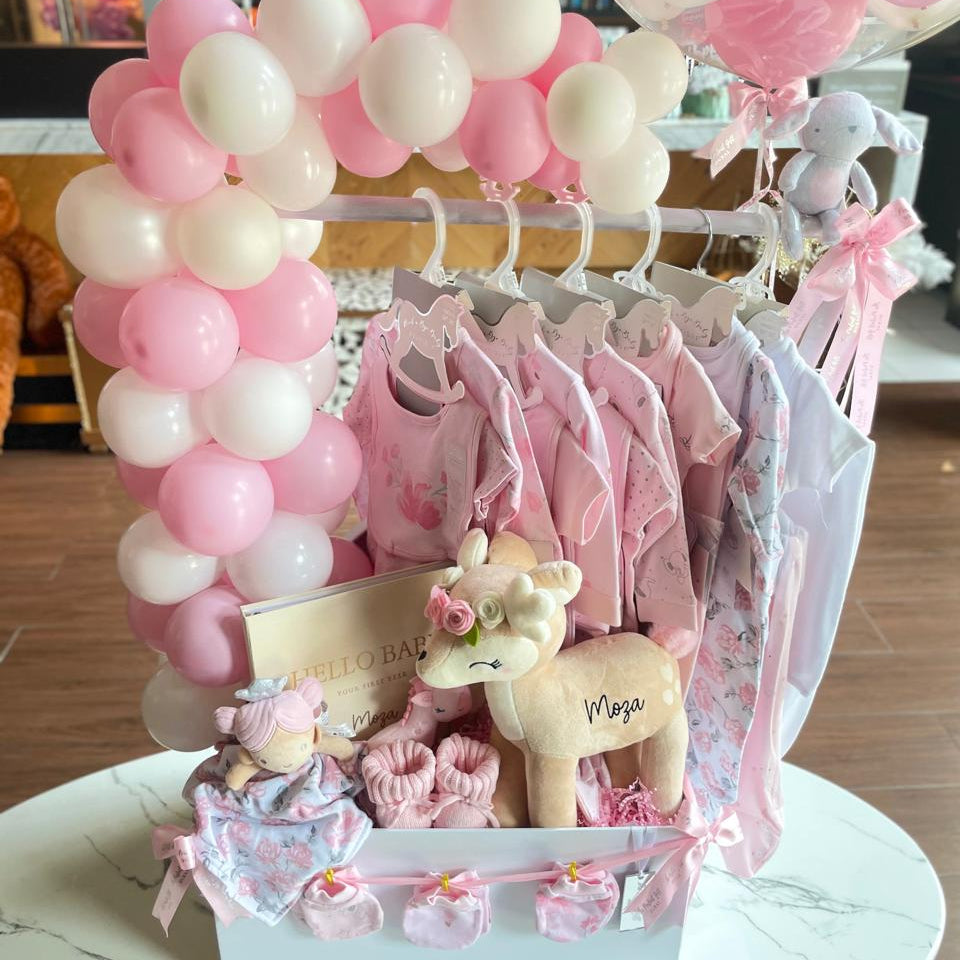 Baby girl gift basket with pink and white balloons, baby clothes, and a toy on a marble surface.