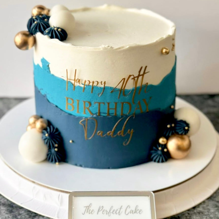 Birthday cake with blue and white design, gold text, and decorative elements on a marble surface.