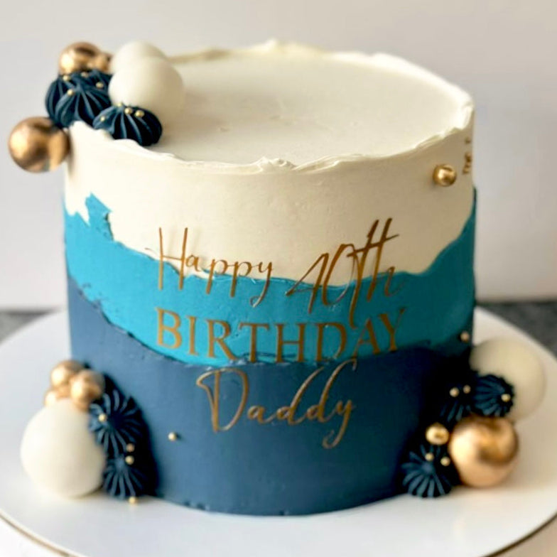 Birthday cake with blue and white design, gold text, and decorative elements on a marble surface.