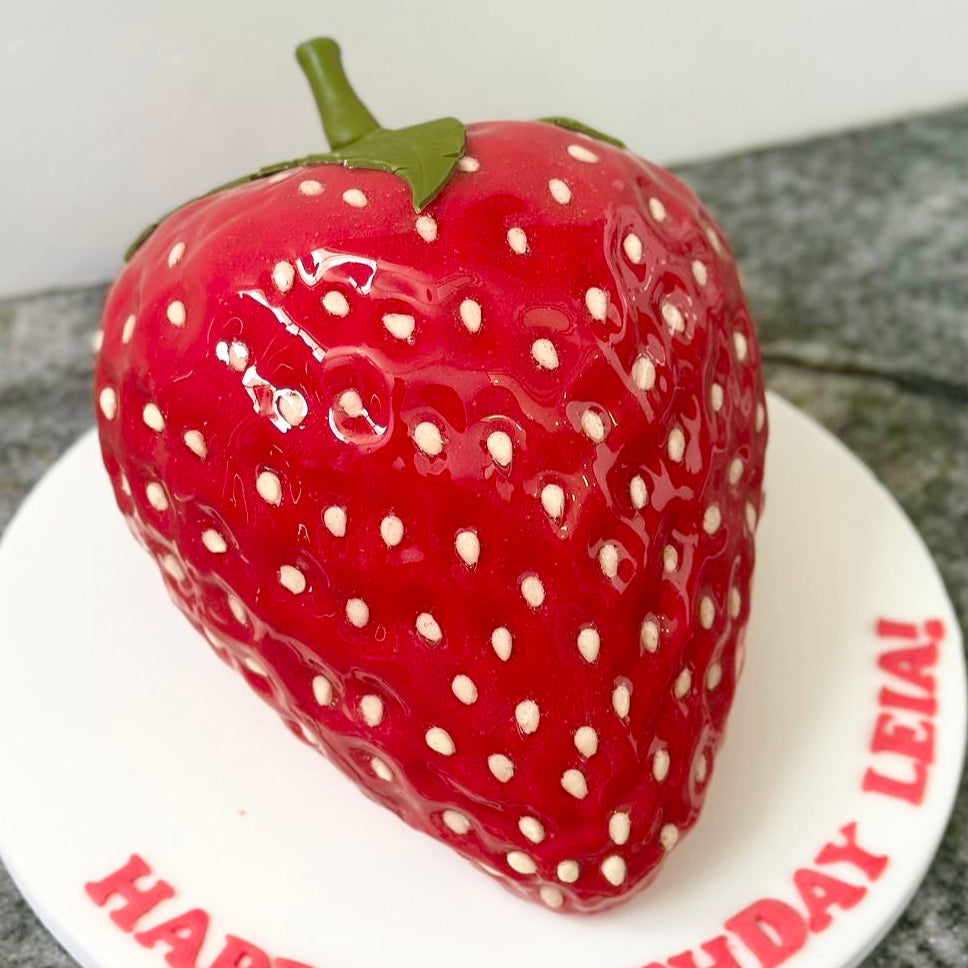 Strawberry-shaped birthday cake with 'Happy Birthday Lelan!' on a white plate.