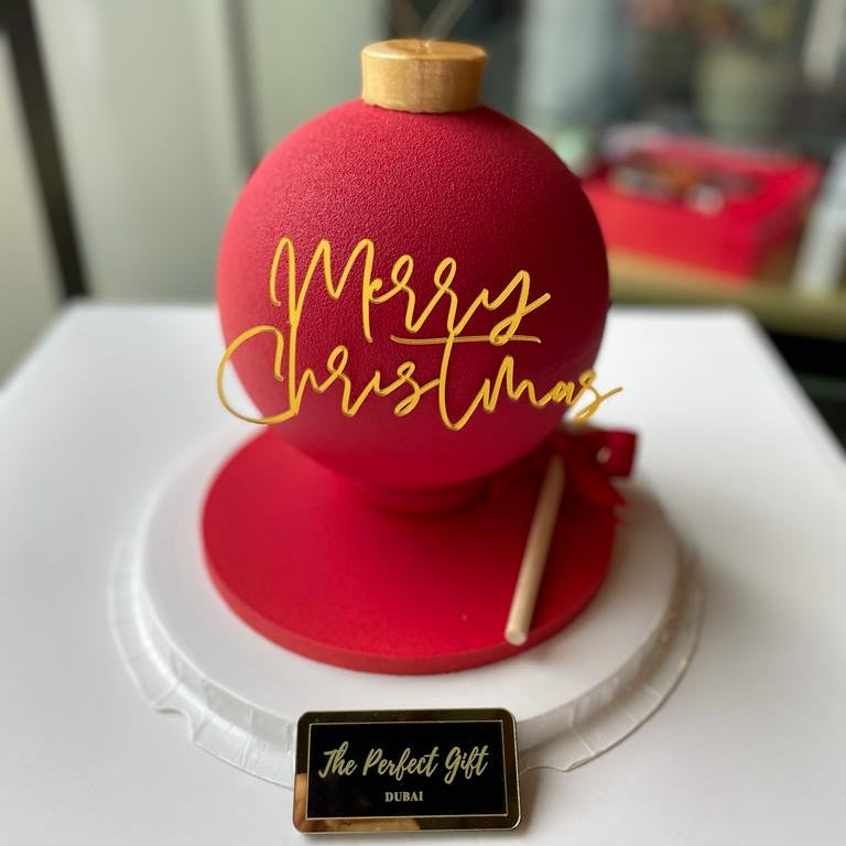 Red Christmas ornament with 'Merry Christmas' in gold on a white surface, branded 'The Perfect Gift'.