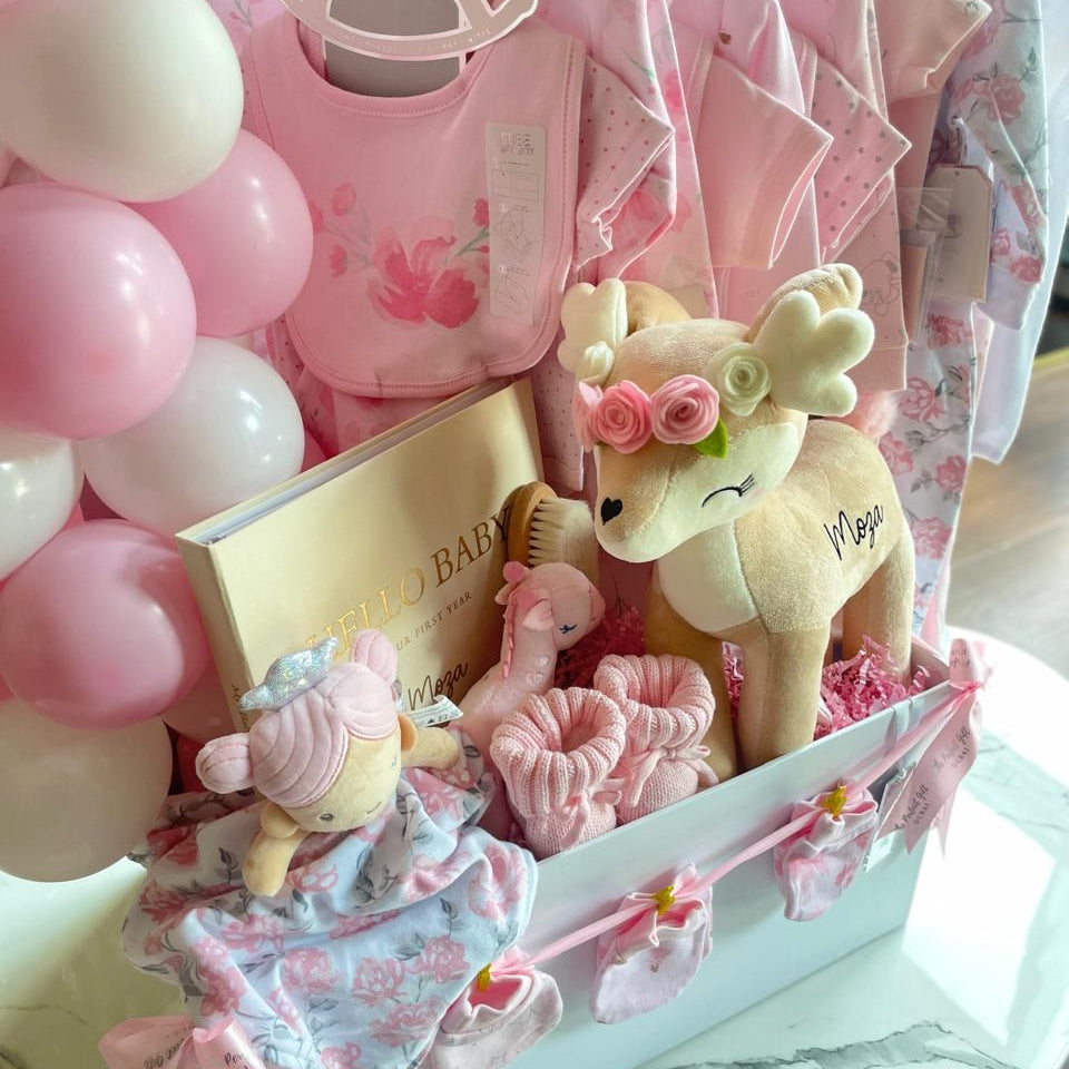 Baby gift basket with pink and white balloons, baby clothes, and a plush toy on a white surface.