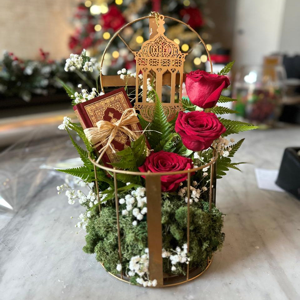 Decorative arrangement with red roses, greenery, and a gold lantern on a marble surface with a blurred festive background.