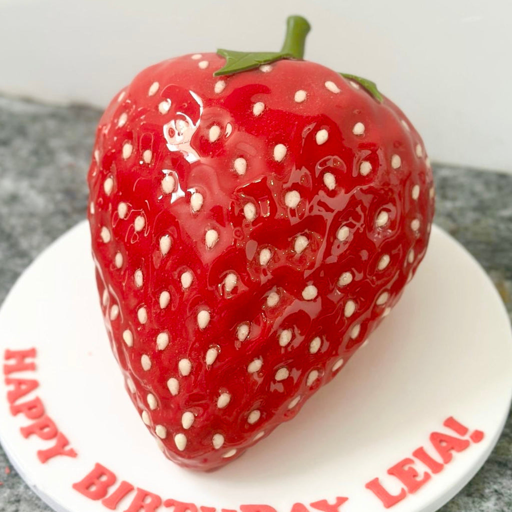 Strawberry-shaped birthday cake with 'Happy Birthday Leia!' text on a white plate.